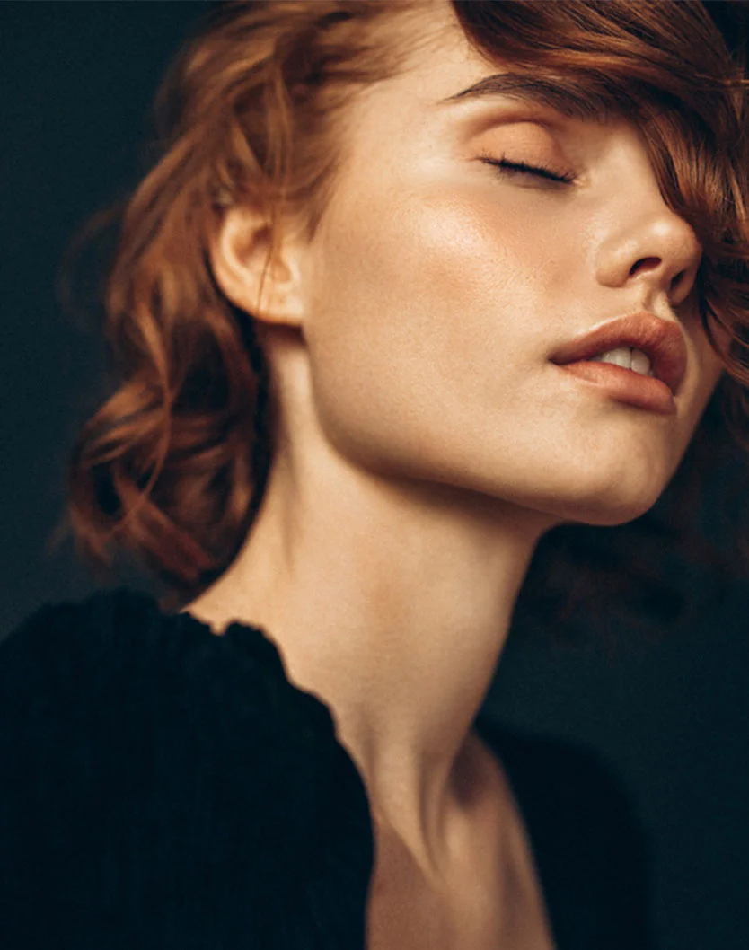 A close-up of a person with short, wavy reddish-brown hair, freckles, and clear skin, leaning their head back with eyes closed, showcasing their jawline and neck. - BBL Therapy in San Fransisco, CA