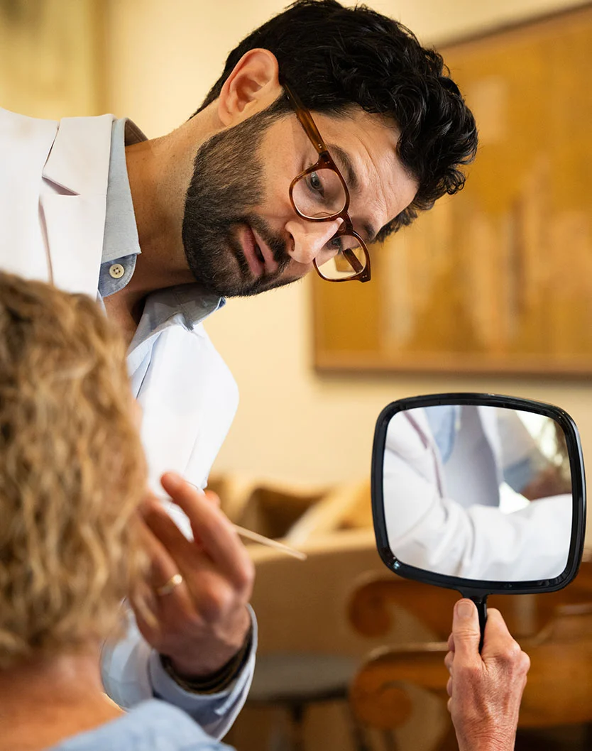 A close-up perspective from over the patient's shoulder, showing a male doctor, Dr. Block-Wheeler, examining the skin of a female patient's face with a cotton swab. The patient is holding a square hand mirror, and the doctor's reflection is visible in the mirror. - Dermabrasion in San Fransisco, CA