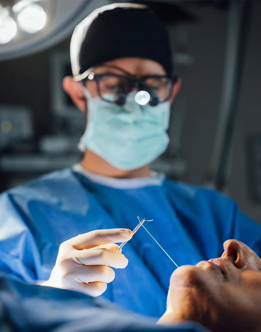 Close-up image of Dr. Block-Wheeler in blue scrubs, a mask, and surgical loupes holding a needle and suture over a patient's mouth/chin area during a procedure. - Facial Skin Cancer MOHs Reconstruction in San Fransisco, CA