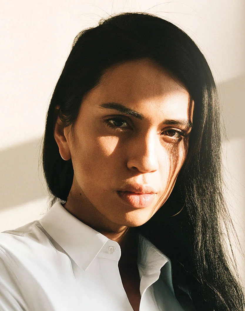 A dramatic, high-contrast portrait of a person with long black hair and a white collared shirt, with strong light casting a sharp shadow across their face. - GA Cheek Implants in San Fransisco, CA