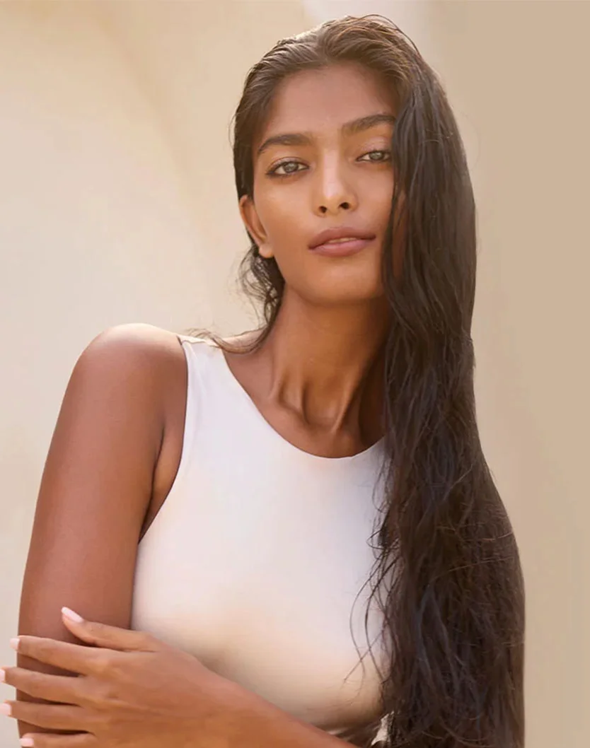 A woman with dark long hair stands in front of a light concrete wall. She holds her arms together under her breasts while wearing a tan one piece swim suit. - Tracheal Shave in San Fransisco, CA
