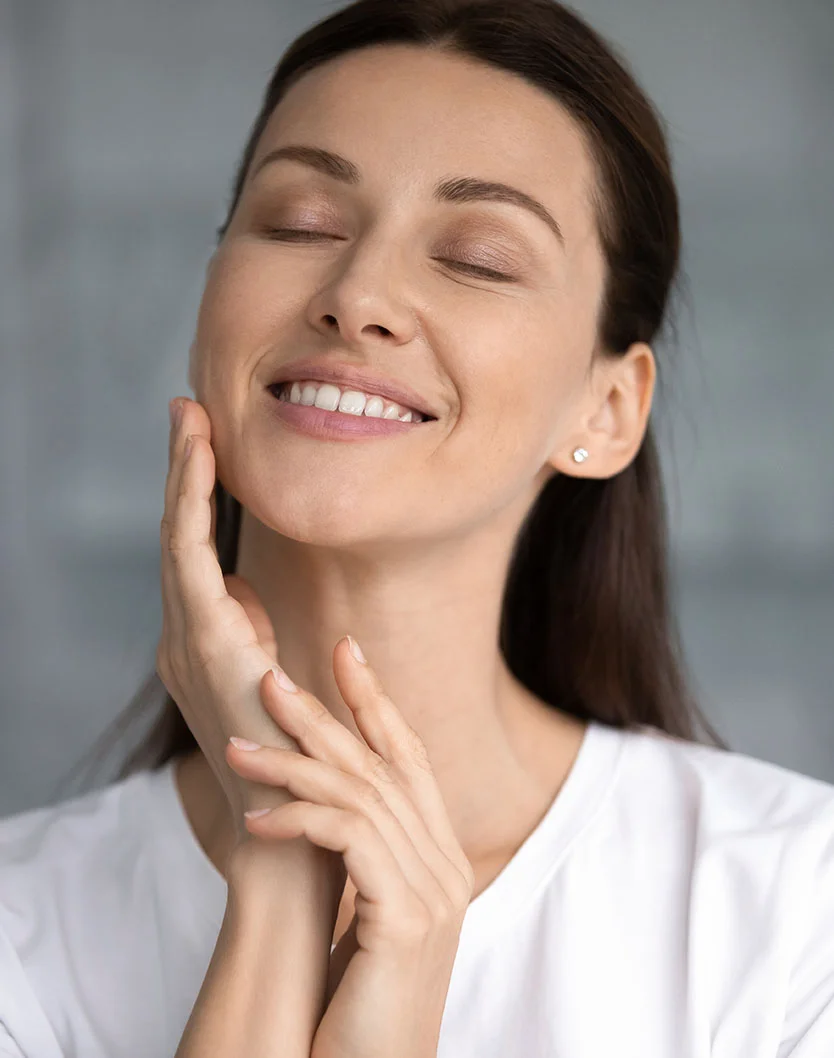 A young woman with long, dark hair looks directly at the camera, gently touching her cheeks with both hands and smiling. She is wearing a white t-shirt, and the background is blurred, showing a light-colored, possibly tiled, surface. - Laser Skin Resurfacing Ablative in San Fransisco, CA