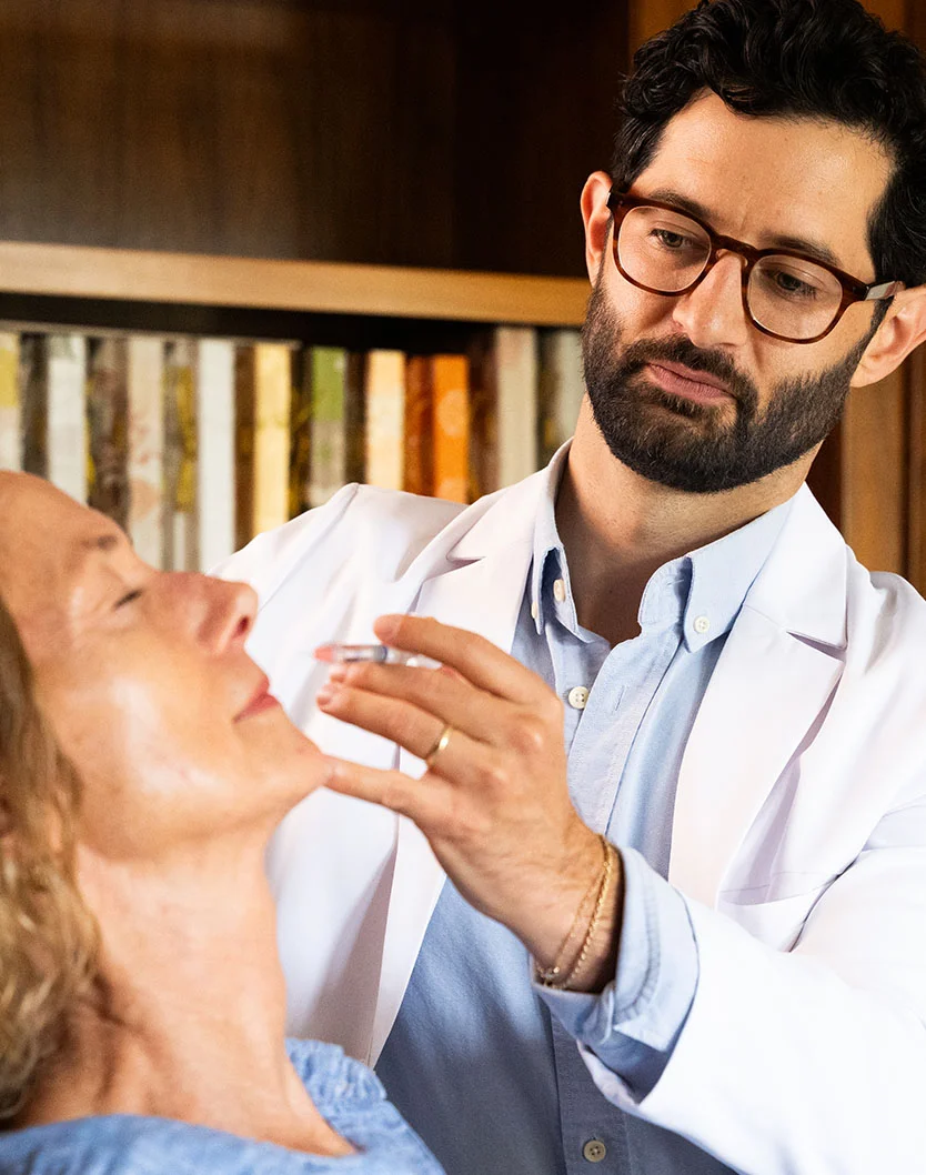 Close-up of a male physician (Dr. Bock-Wheeler) wearing glasses and a white coat, administering an injection (likely a filler) to the lower facial area of an older female patient. - Lip Fillers in San Fransisco, CA