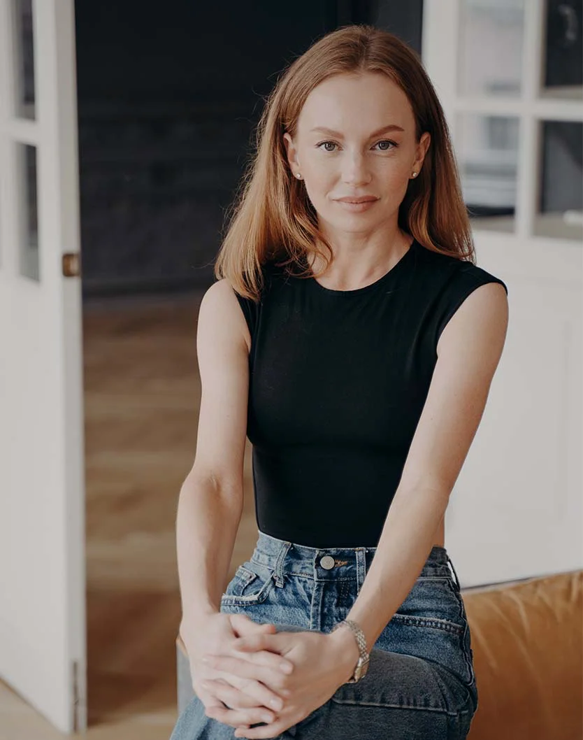 A fair-skinned young woman with long, reddish hair, wearing a black sleeveless top and denim jeans, looking directly at the camera with a slight smile in a bright, indoor setting, illustrating aesthetic outcome. - Lip Reconstruction in San Fransisco, CA