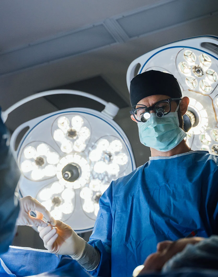 A surgeon (Dr. Bock-Wheeler) wearing a surgical cap, glasses, mask, loupes, and a blue gown, looking down and receiving a surgical tool from an assistant's gloved hand during a procedure, with bright surgical lights visible in the background. - Mole cyst Removal in San Fransisco, CA