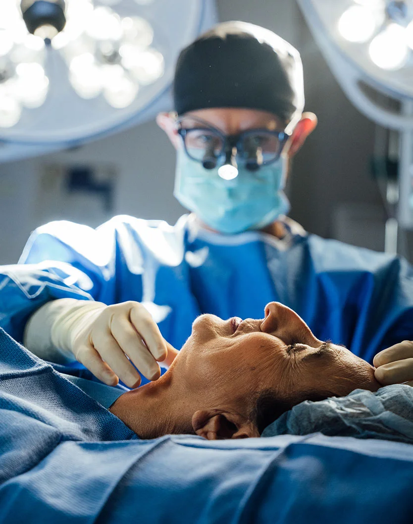 A surgeon (Dr. Bock-Wheeler) wearing a surgical mask, loupes (magnifying glasses), and a blue gown, tending to the neck area of a patient lying on the operating table, with bright surgical lights overhead, suggesting a procedure like neck liposuction. - Neck Liposuction in San Fransisco, CA