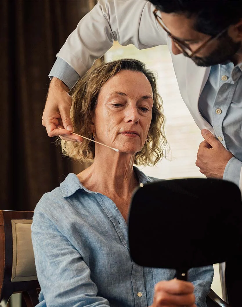 A male physician (Dr. Bock-Wheeler) in a white coat, with his back partially to the camera, using a thin applicator or swab to gently examine or point to the lower jaw and neck area of an older female patient who is holding a handheld mirror. - Neck Lift in San Fransisco, CA