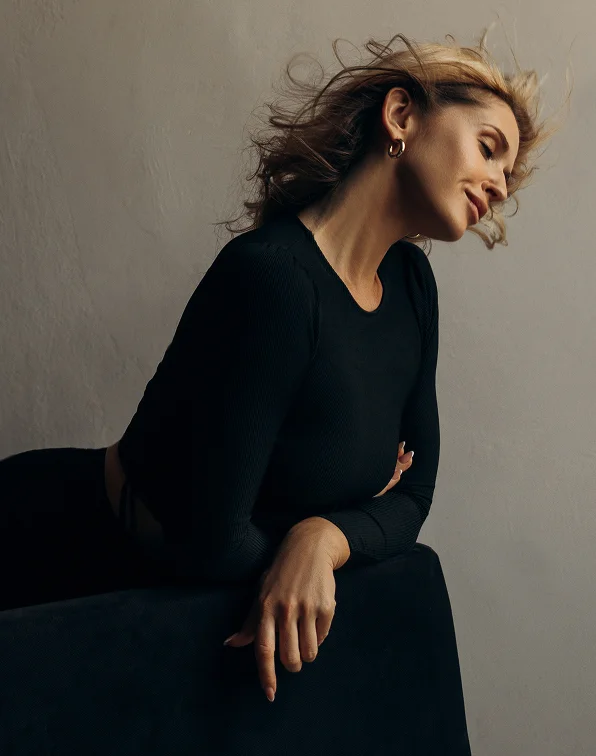 Side profile of a woman with blonde hair wearing a black long-sleeved ribbed top, leaning on a dark velvet surface with her eyes closed, looking serene.