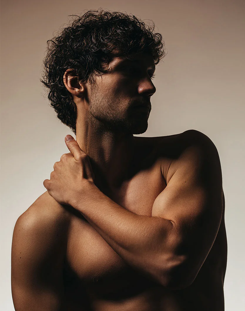 Torsal and profile shot of a shirtless man with dark, curly hair, holding his shoulder, in dramatic lighting. - Laser Hair Removal in San Fransisco, CA